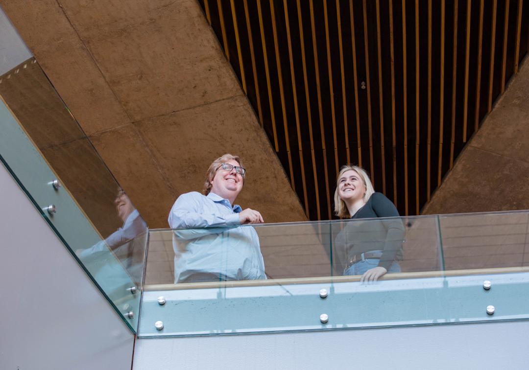 Professor Alastair Blanshard and UQ humanities student, Amelia, stand behind a glass bannister smiling 